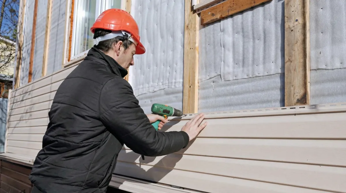 Siding replacement professional using a power drill to install new tan vinyl siding on a home exterior in Dover, DE